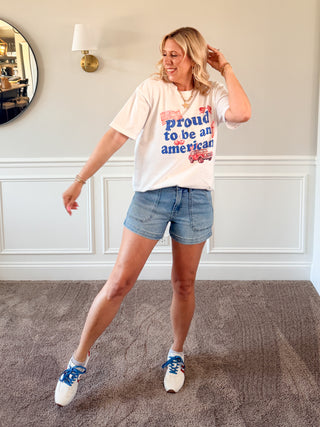 Woman wearing a t-shirt with text Proud to be an american and denim shorts in a room.