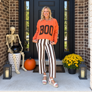 Burnt orange BOO polka dot sweatshirt with striped pants on a Halloween porch with a skeleton, pumpkin and yellow mums - Limeberry Designs